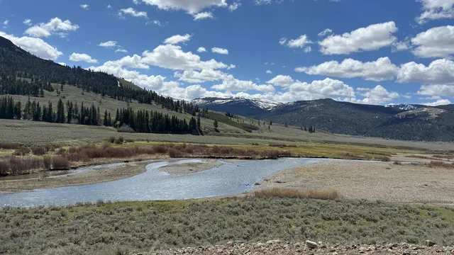 Shallow river winding through a sagebrush valley below forested hills and snowy peaks in Montana
